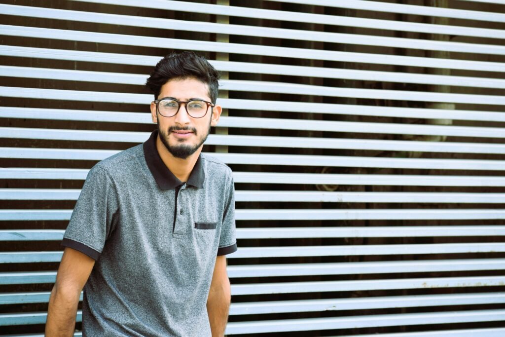 A young man wearing eyeglasses stands against a modern striped wall in casual attire.
