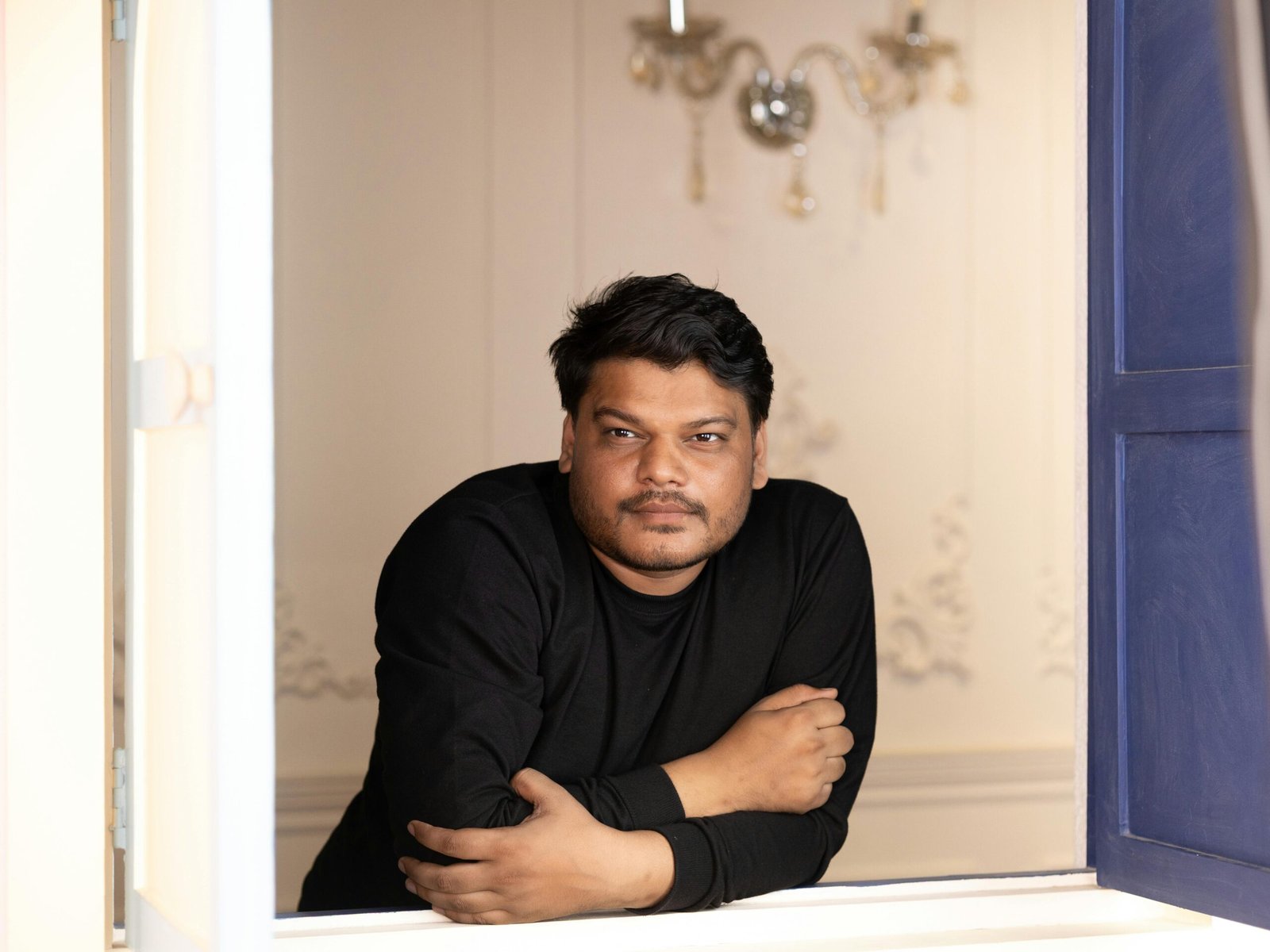 A relaxed man in a black shirt leaning on a window ledge indoors.